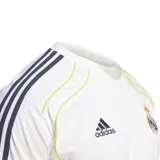 Camiseta de fútbol Adidas del Real Madrid, color blanco con detalles en azul marino y amarillo. Presenta el logo de Adidas en el pecho izquierdo y el escudo del Real Madrid en el pecho derecho. Tiene tres rayas azules en los hombros y cuello redondo. Detalles en amarillo en los laterales y cuello.