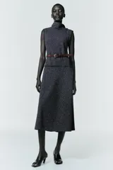 Vestido midi de tejido texturizado en color gris oscuro, con cuello alto fluido, diseño sin mangas y acabados deshilachados. Incluye un cinturón fino de cuero en color marrón con hebilla metálica y cierre de cremallera oculta en la espalda.