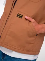 Chaleco de trabajo Caterpillar en color tostado, confeccionado en lona resistente. Presenta cierre frontal completo, cuello tipo bomber en tejido acanalado, bolsillos frontales tipo parche y etiqueta de la marca en el pecho.