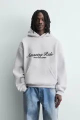 Sudadera con capucha y bolsillo frontal tipo canguro, confeccionada en tejido de algodón con interior perchado. Presenta diseño de corte boxy fit, mangas largas con puños y ruedo en rib, y texto bordado y estampado en el frente.