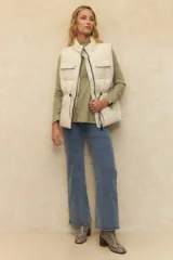 Chaleco puffer color crema, con cuello alto, cierre frontal y cuatro bolsillos con solapa. Cuenta con cintura ajustable con cordón.