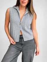 Chaleco de punto trenzado color gris, sin mangas, con cuello alto y cierre frontal doble. Presenta un diseño de tejido con textura de ochos y terminaciones acanaladas.