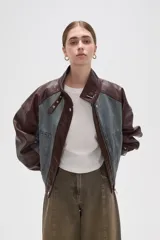 Campera estilo bomber con diseño color block, combinando paneles de denim lavado con mangas y detalles en cuero sintético color marrón oscuro. Presenta cuello alto con tira ajustable, cierre frontal con botones a presión y bolsillos frontales tipo parche.