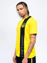 Remera amarilla con una franja vertical negra en el lado izquierdo. En la parte superior de la franja se encuentra el escudo del Club Atlético Peñarol. En la parte inferior de la franja se lee la leyenda "Oro y Carbón".