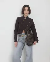 Campera de estilo militar en color marrón oscuro, confeccionada en gamuza. Presenta un diseño corto, cuello alto y cierre frontal cruzado con botones metálicos decorativos. Incluye detalles de charreteras en los hombros y botones en los puños.