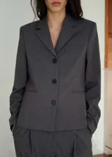 Chaqueta de corte recto estilo sastre, color gris, con cuello de solapa con muesca, cierre frontal de tres botones, bolsillos laterales de ribete y un bolsillo de ribete en el pecho. Cuenta con forro interior y mangas largas.