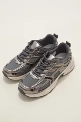 Championes urbanos de estilo deportivo, confeccionados en una combinación de malla técnica gris y detalles en cuero sintético metalizado color peltre. Presentan cordones al tono, suela de goma con amortiguación en color crema y diseño de paneles superpuestos.