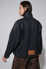 Campera negra oversized estilo utilitario con botones metálicos, hombros caídos, bolsillos con solapa y terminación levemente fruncida en el ruedo. El acabado tipo cuero aporta textura y un carácter urbano distintivo.