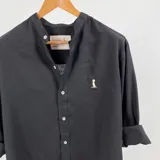 Camisa negra de manga larga con cuello mao y botones frontales blancos. Presenta un pequeño logo bordado en color arena en el pecho izquierdo.