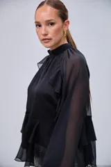 Blusa negra de cuello alto con volados en el pecho y mangas largas con transparencias. Presenta un diseño escalonado con volados en la parte inferior.