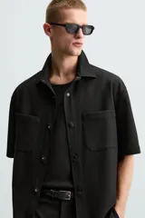 Sobrecamisa negra de tejido con elasticidad, cuello solapa y manga corta. Tiene dos bolsillos de plastrón en el pecho y cierre frontal con botones a presión.
