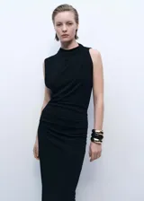 Vestido largo negro de diseño recto y sin mangas, confeccionado en tejido elástico. Presenta cuello drapeado y detalle de drapeado en la cintura, con una abertura lateral.
