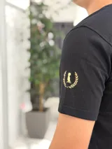 Remera color beige con cuello redondo y mangas cortas. Estampado frontal con el texto "Duxonstudios" en tipografía cursiva, acompañado de un logotipo circular con hojas de laurel y una figura central en tono verde oscuro.