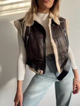 Chaleco marrón de cuero sintético con interior de corderito color crema, cierre frontal con cremallera, bolsillos laterales con cremallera y cinturón con hebilla.