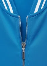 Chaqueta tipo bomber color turquesa con cierre frontal de cremallera, cuello redondo y puños con detalles de rayas blancas en contraste. Presenta un diseño recto con dos bolsillos laterales.