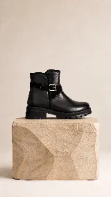 Bota corta de cuero negro con suela track de goma, detalle de tira lateral con hebilla metálica y acabado minimalista.