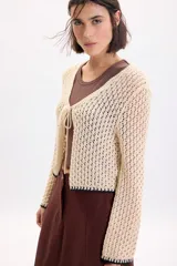 Cardigan de punto fantasía color crudo, con mangas largas y cierre delantero con tiras para atar. Presenta detalles de terminación en color negro en el ruedo y los puños.