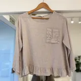 Blusa beige de manga larga, con escote redondo, bolsillo frontal con aplique bordado y detalle de volados en el ruedo.
