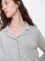 Camisa de pijama de manga larga con cuello solapa y cierre frontal de botones. Presenta un estampado sutil de pequeños corazones sobre una base de color gris melange, con detalles de ribete en contraste en el cuello y la botonera.