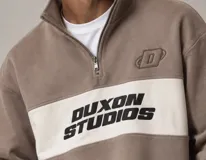 Buzo de cuello alto con medio cierre y diseño de bloques de color marrón claro y una franja central blanca. Presenta el nombre de la marca 'Duxon Studios' estampado en negro en el frente y un logo bordado en el pecho.
