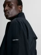 Campera negra de nailon arrugado reciclado, con cuello alto, puños y bajo elásticos, cierre con cremallera, bolsillos con cremallera en la cintura, abertura de ventilación en la espalda, forro de poliéster reciclado e insignia con el logo de goma de Calvin Klein en la manga.