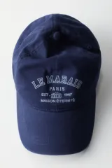 Gorra de sarga de algodón en color azul marino con efecto lavado, visera curva y diseño bordado en el frente con el texto 'Le Marais Paris Est. 1947 Maison Éternité'. Cuenta con botón superior y trabilla ajustable con cierre de metal en la parte trasera.