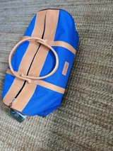 Bolso de viaje tipo duffel de lona azul con detalles y asas de cuero tostado, cierre superior y diseño espacioso.