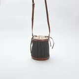 Bolso tipo saco con cuerpo de tela a rayas verticales en blanco y negro, con detalles de cuero sintético color marrón en la base, borde superior y correa ajustable.