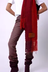 Bufanda de lana merino tejida en telar, con diseño de rayas en tonos marrón, beige y rojo. Presenta flecos en los extremos y un parche negro con la palabra "CAPER" en blanco.