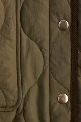 Campera de tejido guateado color verde oliva con ribetes de pana en contraste. Presenta cuello redondo, cierre frontal con botones a presión, bolsillos delanteros de parche y bajo recto con aberturas laterales redondeadas.