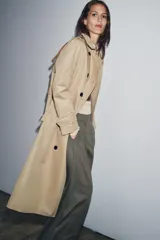 Trench color beige con cuello solapa y manga larga ajustable con trabillas. Bolsillos en delantero. Cinturón en mismo tejido. Detalle de pespuntes marcados a tono. Cierre frontal cruzado con botones. Tejido repelente al agua.