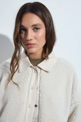 Campera de corderito color beige, con cuello camisero y cierre frontal de botones a presión. Presenta una cartera central lisa en contraste y mangas largas con puños elásticos.