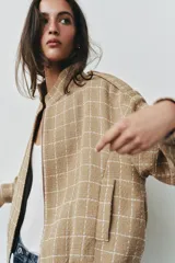 Chaqueta bomber de color beige con diseño a cuadros blancos, cuello subido, manga larga con trabillas en los hombros, bolsillos delanteros y bajo con elástico. Cierre frontal con cremallera y botones a presión.