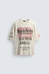 Camiseta de punto de algodón con corte boxy fit, cuello redondo y manga corta. Presenta un estampado frontal con temática de taller mecánico en tonos rojo desgastado y negro, incluyendo el texto "Rust & Revive", "Old Iron Garage" y detalles técnicos. La prenda tiene un efecto lavado y acabados irregulares.