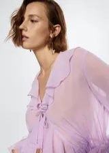 Vestido largo de tejido fluido y semitransparente color lila, con cuello en pico, mangas largas con volantes y cierre frontal con lazo. Presenta volantes decorativos a lo largo de la abertura frontal y fruncido elástico en la cintura.