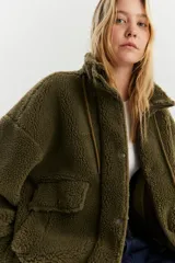 Campera de sherpa color verde musgo, con cuello alto, cierre frontal de cremallera y solapa con botones a presión. Presenta bolsillos delanteros con solapa y cordón de ajuste en el ruedo.