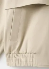Chaqueta oversize de algodón color beige, con cuello alto, cierre frontal oculto con cremallera, mangas largas con puños abotonados, bolsillos laterales y bajo fruncido elástico.