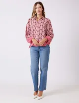 Camisa de gasa con cuello con solapa y apertura central con botones. Presenta un estampado de patrones geométricos en tonos rosados, marrones y crema, con un borde inferior en un patrón geométrico más denso en tonos rojizos/rosados. Las mangas son amplias de largo tres cuartos con puño elastizado.