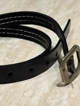 Cinturón fino de cuero negro con pespuntes en contraste y hebilla metálica de diseño hexagonal en tono bronce envejecido.