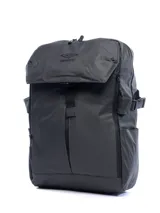Mochila de color gris oscuro o negro, con acabado liso y mate, de estilo urbano y cierre superior tipo solapa con hebilla ajustable. Presenta el logo de Umbro bordado en la solapa.