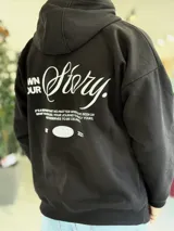 Canguro oversize negro con capucha y bolsillo tipo canguro. Presenta un gran estampado en la espalda con la inscripción "Own Your Story", texto descriptivo y un gráfico de globo terráqueo junto al año "2021".