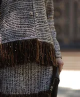 Chaqueta de tweed en tonos beige y marrón, con cuello redondo, cierre frontal con botones joya y detalle de flecos largos en el ruedo.