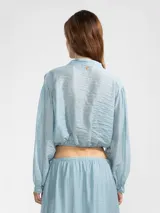 Blusa corta de tejido arrugado liviano, con cuello camisero, cierre frontal de botones y mangas largas abullonadas con puños. Presenta un ruedo elastizado que le otorga una silueta amplia y relajada.