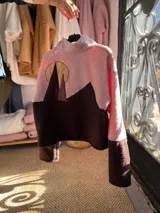 Buzo de polar tipo corderito con diseño de bloques de color en tonos rosa y marrón chocolate. Presenta cuello alto con cierre frontal parcial y un aplique circular en el pecho.