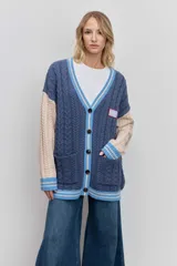 Cardigan de punto con diseño de bloques de color, combinando azul marino en el cuerpo y beige en las mangas. Presenta un tejido con textura de trenzas, cuello en V con borde rayado en azul y blanco, cierre frontal con botones y bolsillos delanteros. Incluye un parche decorativo en el pecho.