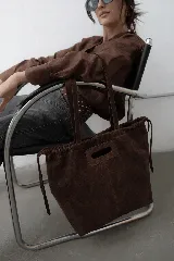 Bolso tipo tote de gamuza color marrón, con diseño de frunces en la parte superior y cierre mediante cordones laterales. Cuenta con doble asa larga para llevar al hombro y una abertura central reforzada para llevar en la mano.