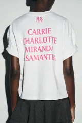 Camiseta blanca de manga corta y cuello redondo, con corte regular y bajo recto. Presenta el logo de "Sex & The City" estampado en color fucsia en el frente.