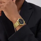 Reloj Guess con caja redonda dorada y esfera negra. Presenta índices dorados y un pequeño cristal en la posición de las 12. La correa es de malla metálica dorada.