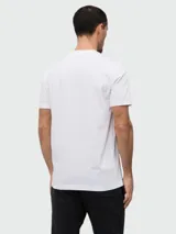 Remera de algodón blanca con cuello redondo y manga corta. Presenta un estampado frontal con el logo de la marca en efecto distorsionado y un parche circular con el logo en la manga.
