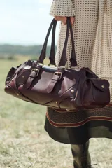 Bolso de hombro alargado de piel color borgoña con doble asa, detalles y hebillas metálicas doradas, charm de espejo colgante y bolsillos laterales exteriores.